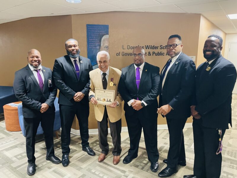 Governor Wilder receives his 75th service medallion and certificate from Omega Psi Phi. Fraternity, Inc. Standing are six men with Governor Wilder in the middle